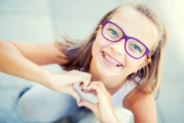 A young girl with blonde hair and glasses smiles to show off her stylish braces.